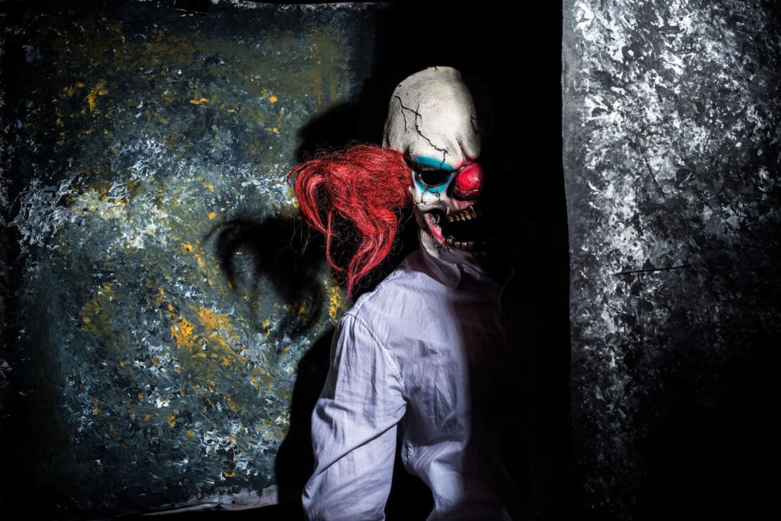 A live actor dressed as a clown peers behind a dark wall, waiting to scare someone