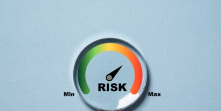 A person holds a magnifying glass. Inside the glass is a scale. The left side of the scale is green with the words 'Min' and the right side is red with the word 'Max.' At the center of the scale is the word 'Risk.'